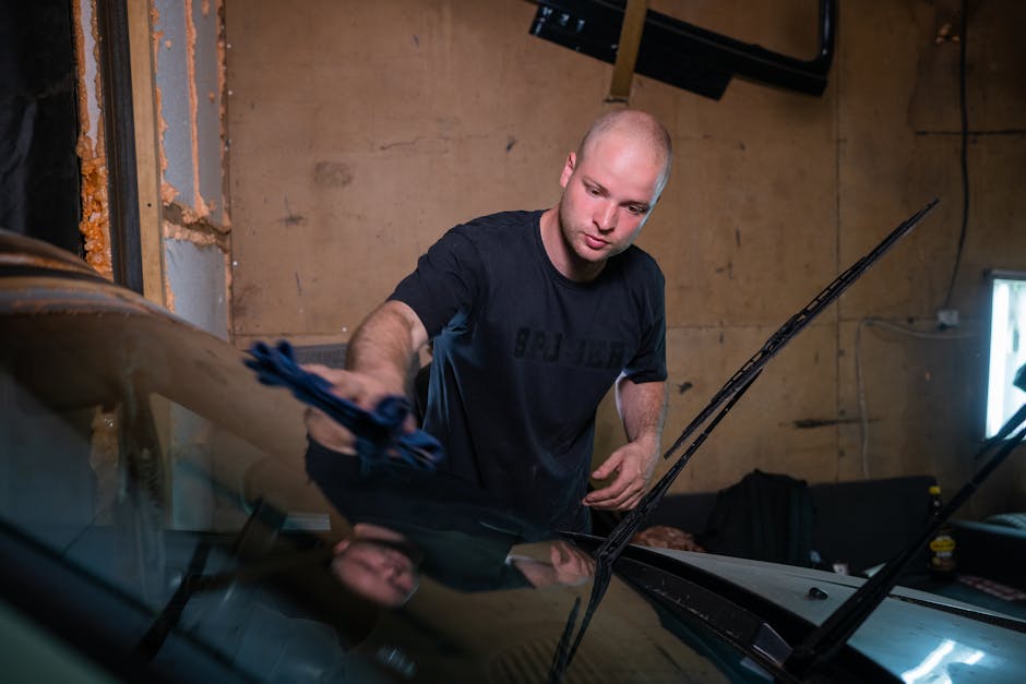 A man wipes a car windshield inside a garage, focused on cleaning.