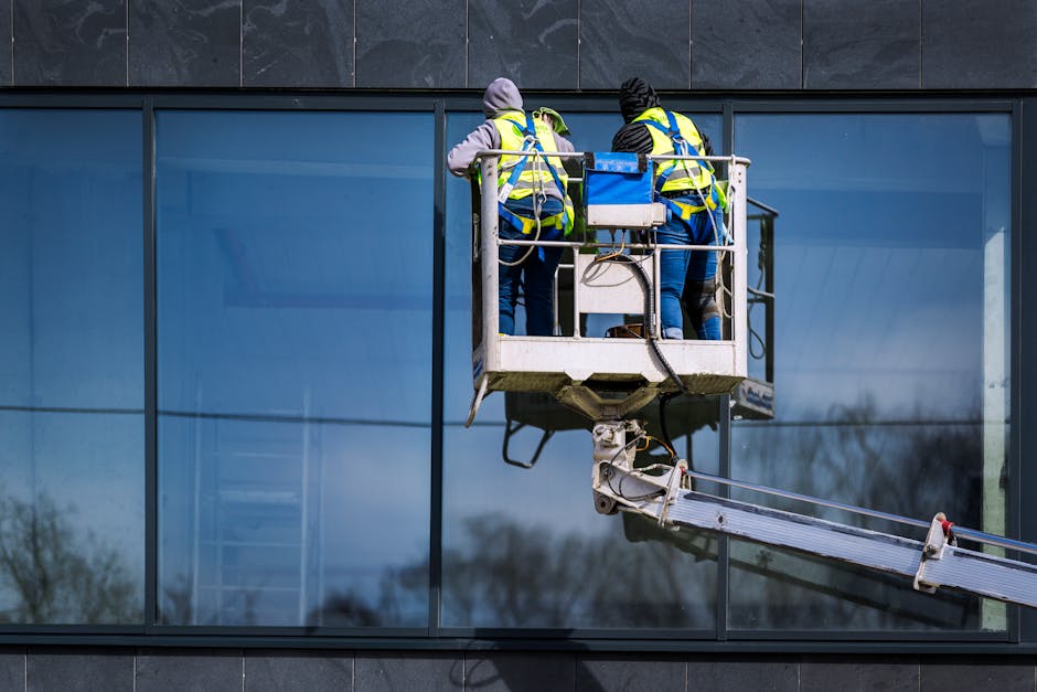 Two men on a hydraulic lift cleaning windows of a modern skyscraper. Urban reflection visible in the glass.