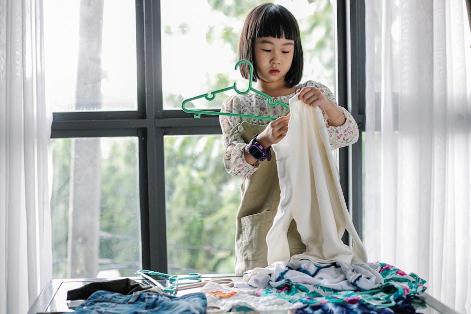 A young girl helping with laundry, folding clothes indoors by a window.